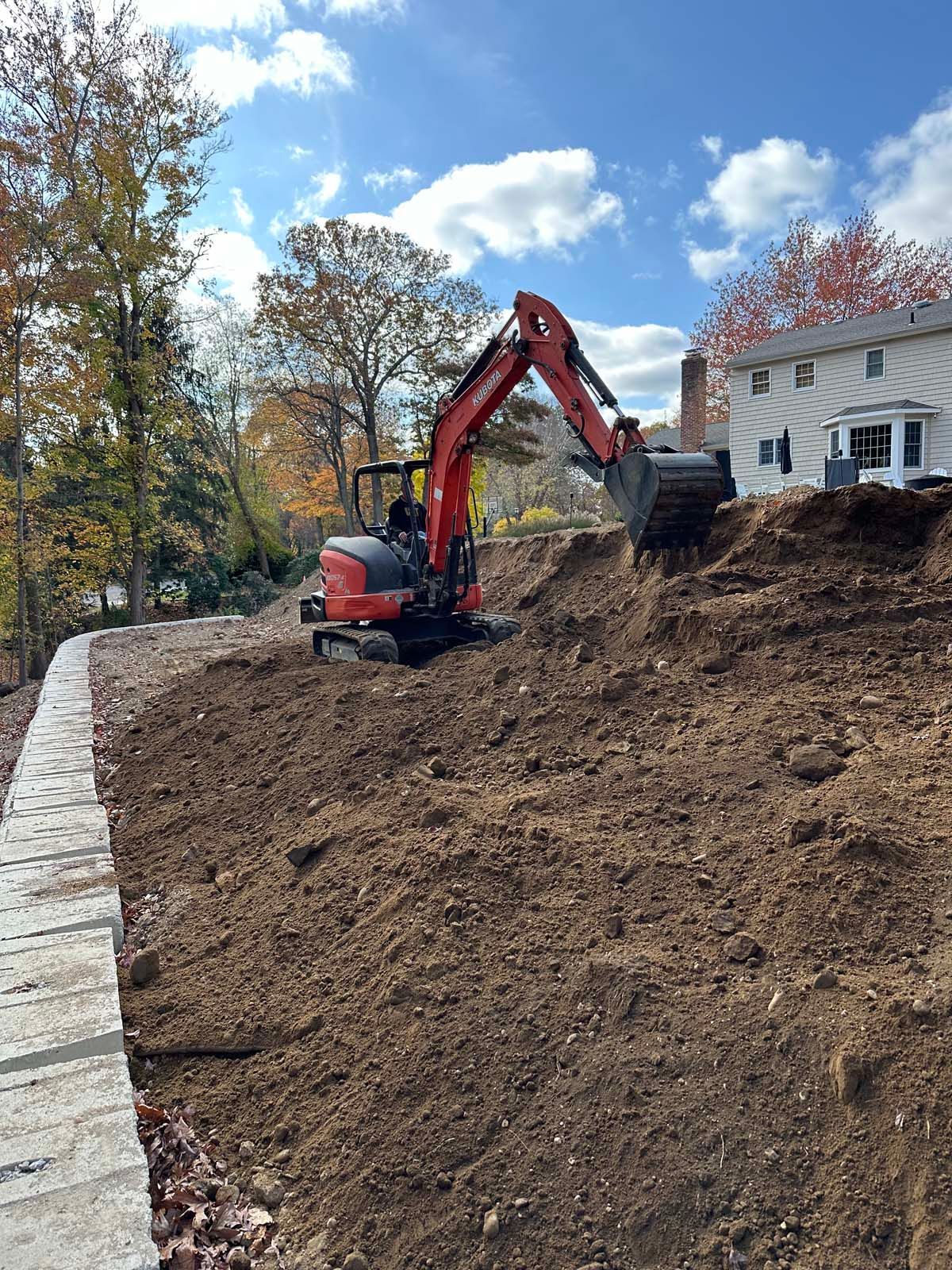 Mini excavator digging dirt near a retaining wall and house under a blue sky.