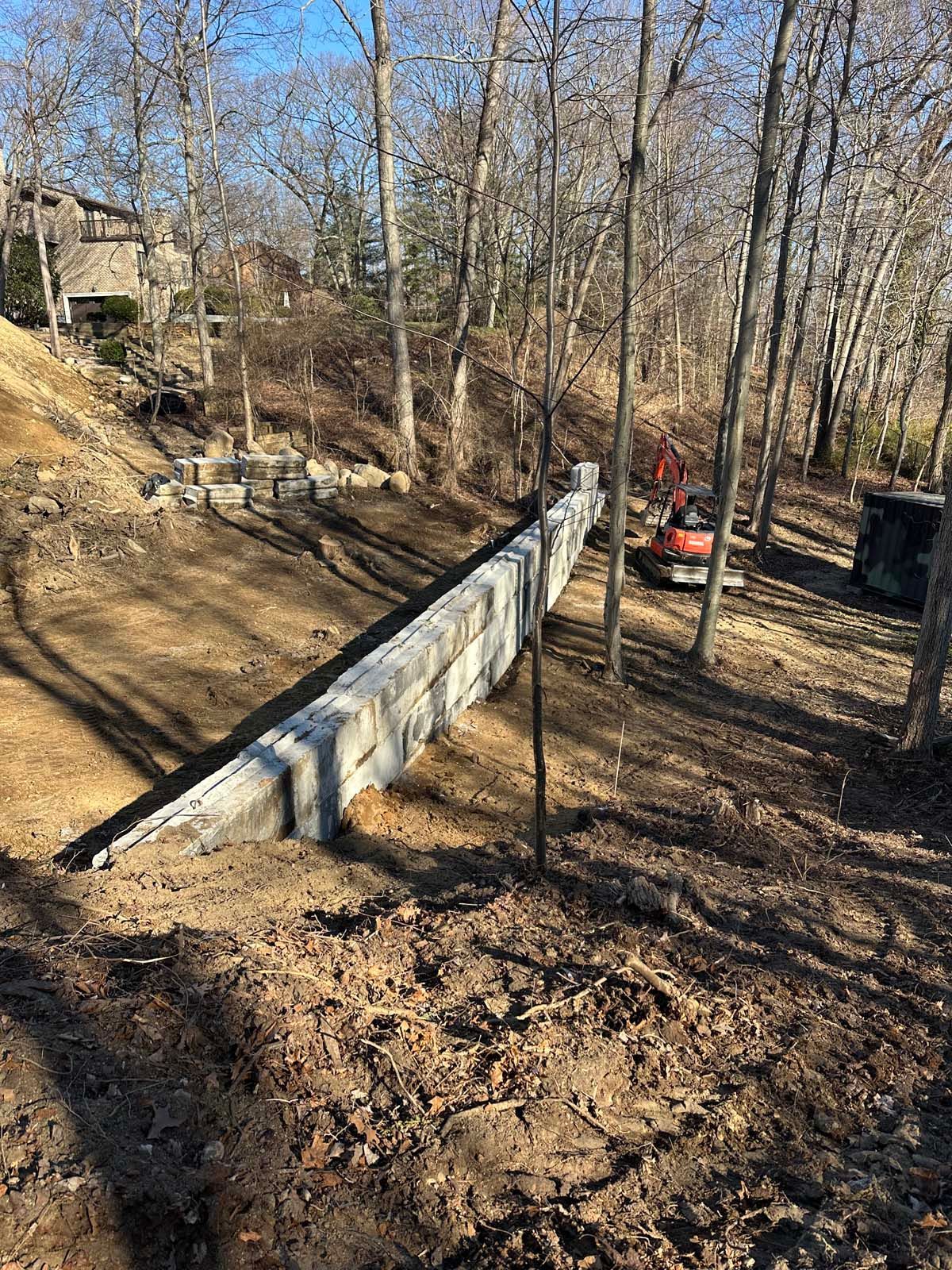 A long, concrete retaining wall under construction in a wooded area; an excavator sits in the background.