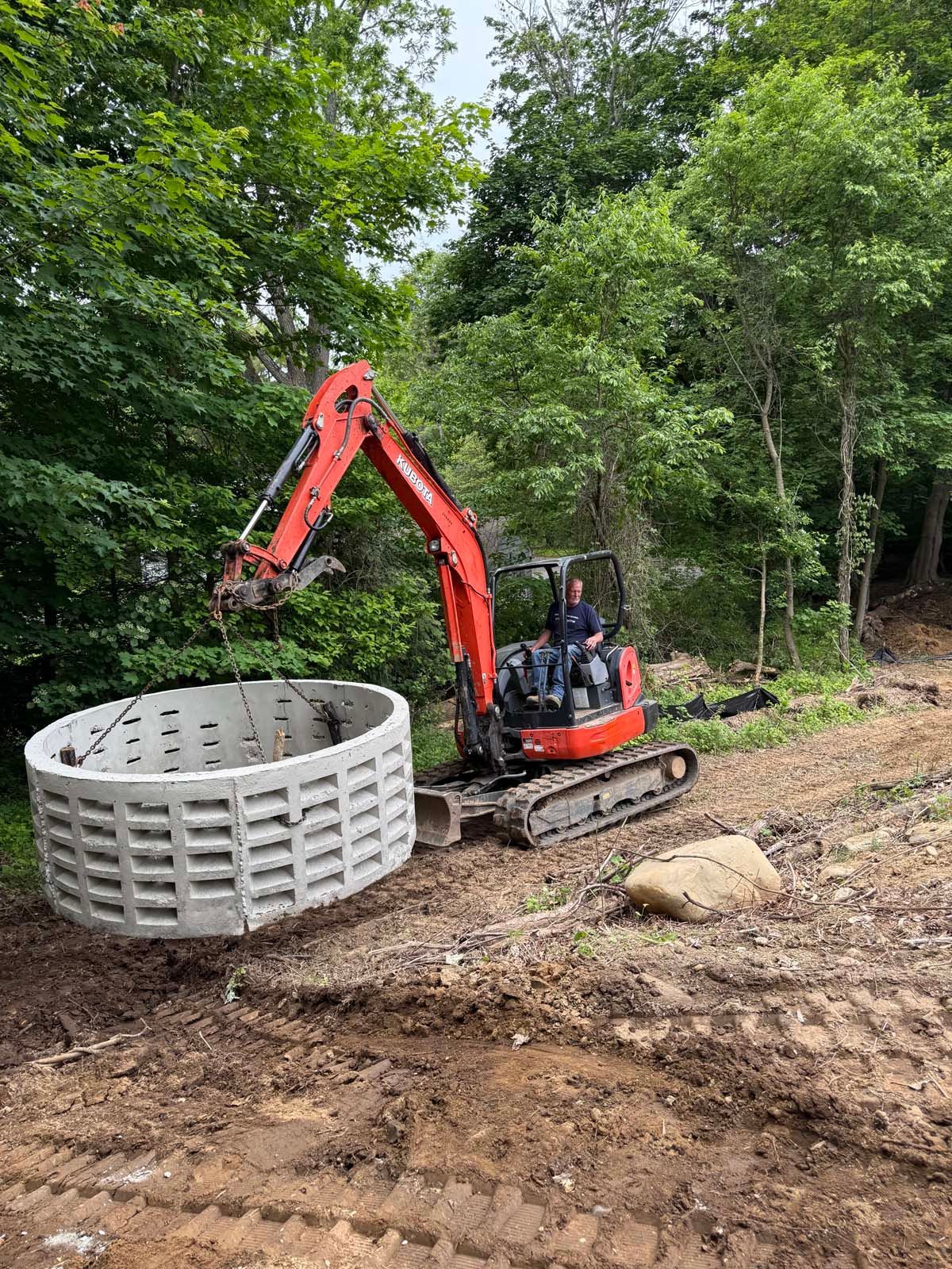 Red excavator lifting a concrete well ring in a wooded area. A person is in the excavator.
