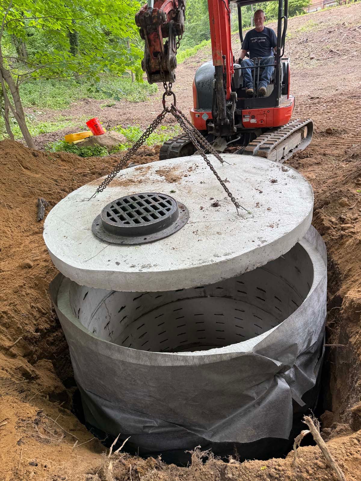 A concrete septic tank cover being lowered onto the tank by an excavator in a dirt trench.