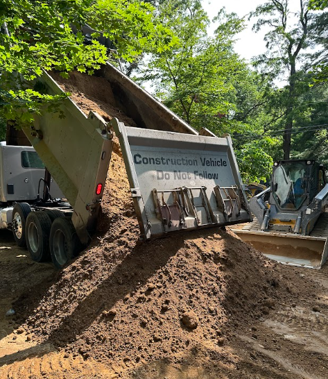 Dump truck unloading soil onto a pile near a construction site, with a skid steer loader visible.