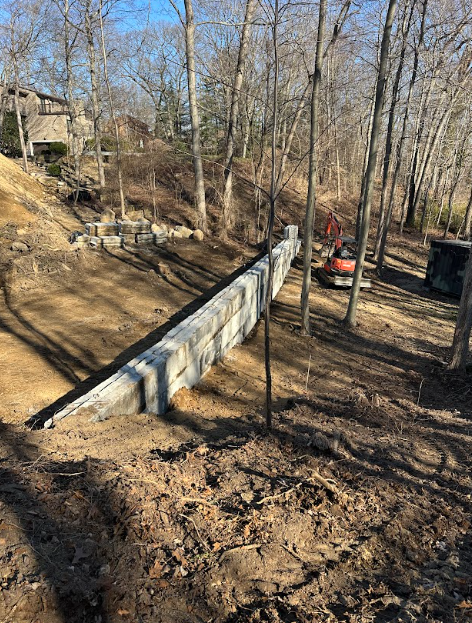 Concrete retaining wall under construction on a hillside, with trees and an excavator nearby.