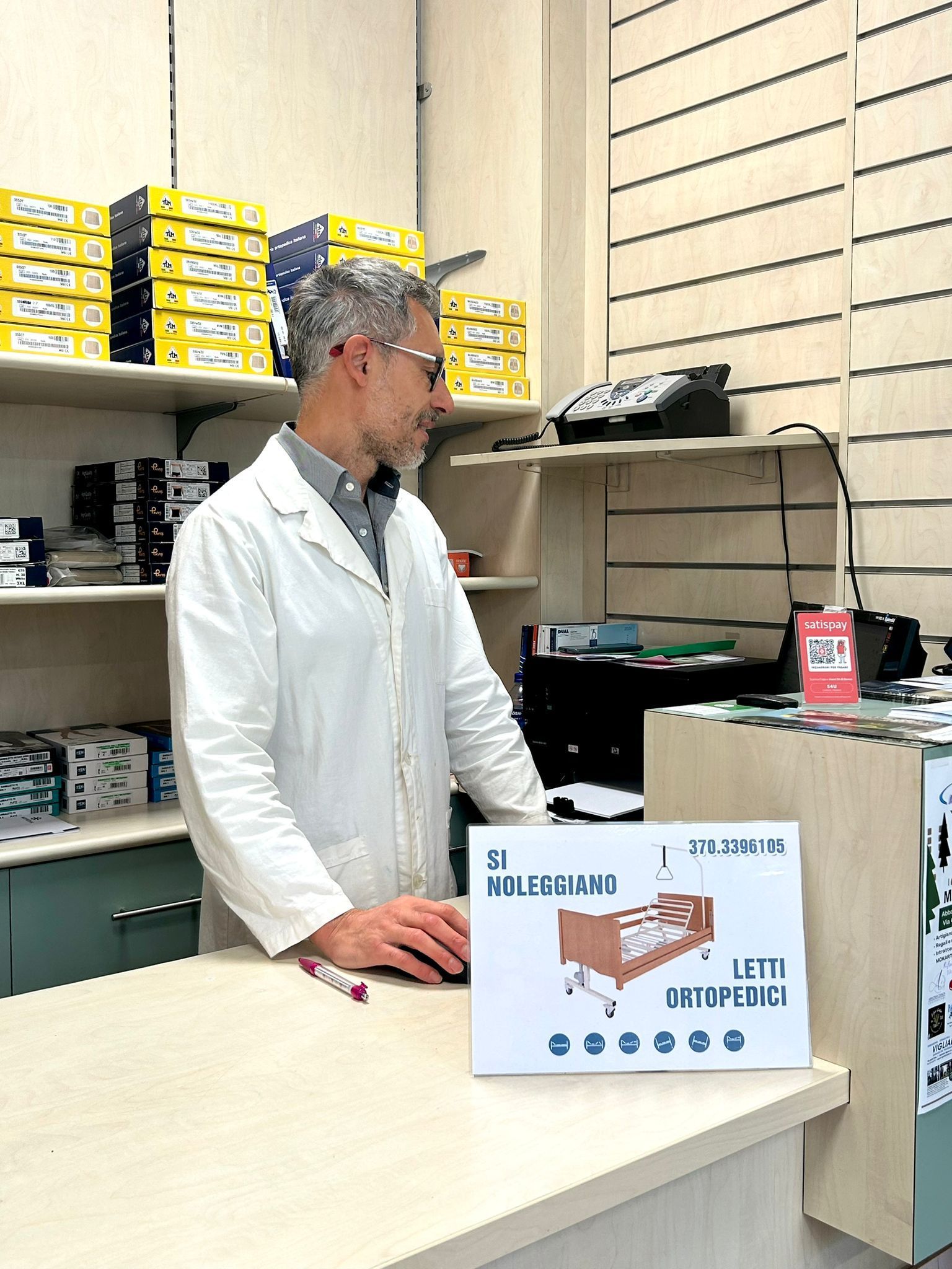 Un uomo in camice bianco è in piedi davanti al bancone di un negozio e guarda un cartello; dietro di lui ci sono scaffali pieni di provviste.