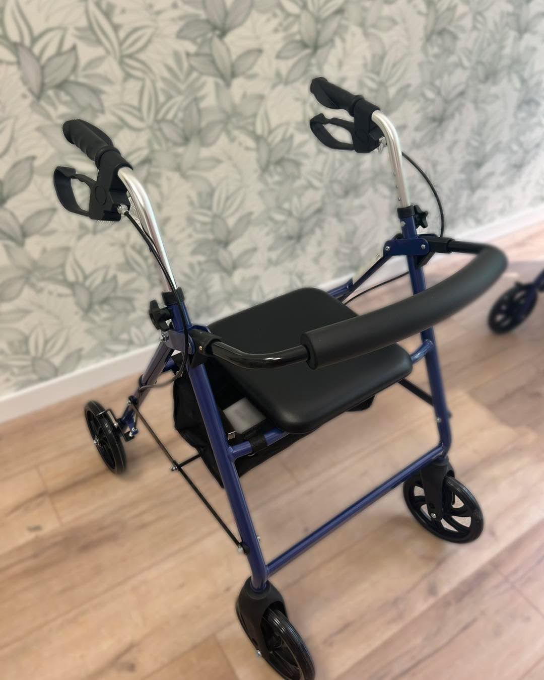 Deambulatore blu con sedile, braccioli e ruote neri, su un pavimento in legno di fronte a una parete decorata.