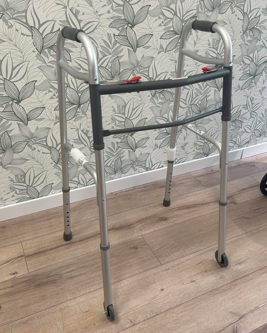 Deambulatore argentato con ruote, altezza regolabile, su un pavimento in legno vicino a carta da parati a motivi.