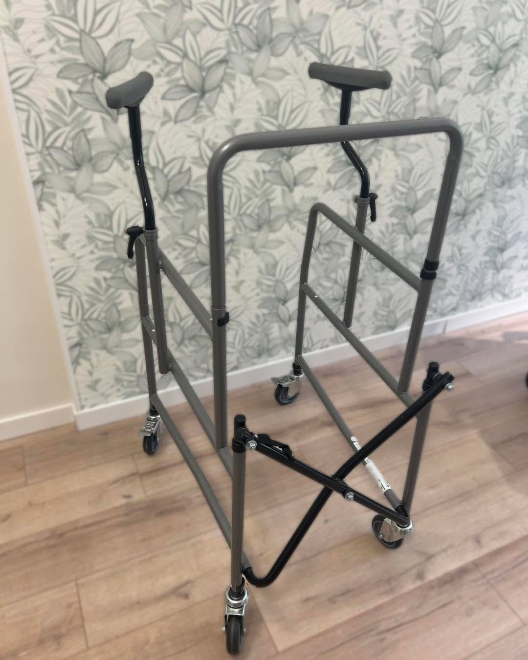 Deambulatore grigio con ruote e braccioli, ripiegato. Appoggiato a una parete con carta da parati floreale e pavimento in legno.