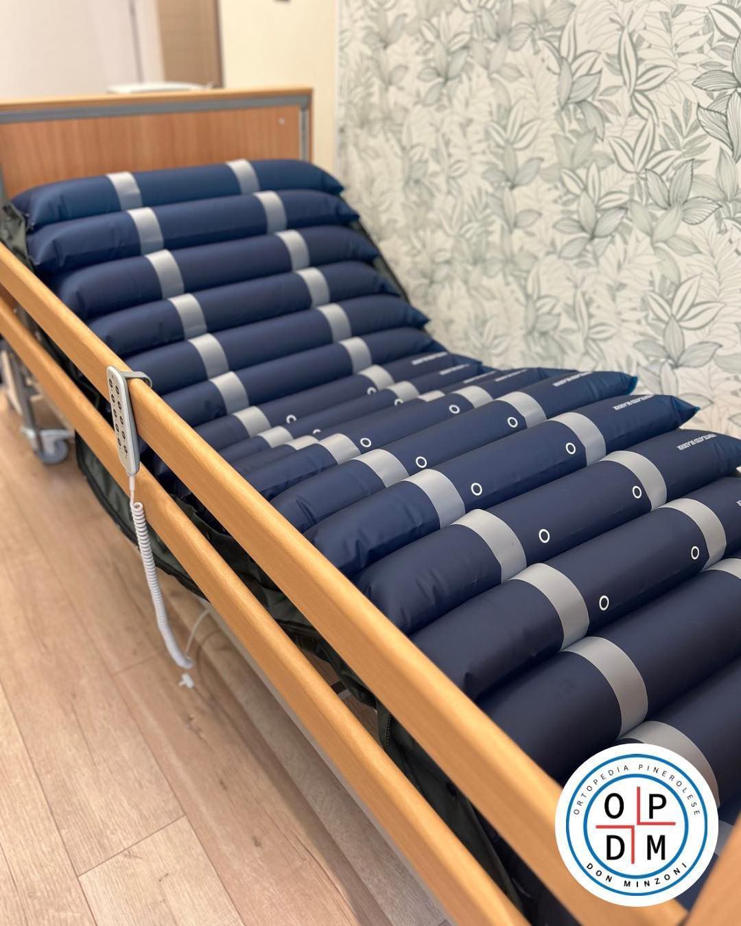 Materasso ad aria blu su una struttura in legno di un letto d'ospedale, in una stanza con carta da parati a motivi.