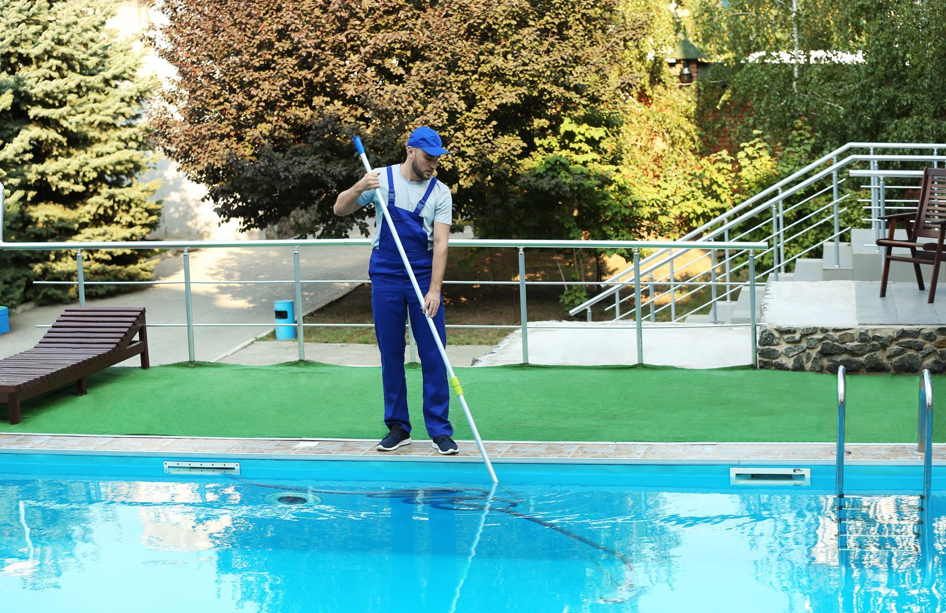 Man in blue overalls cleaning a pool with a long-handled net, sunny outdoor setting.