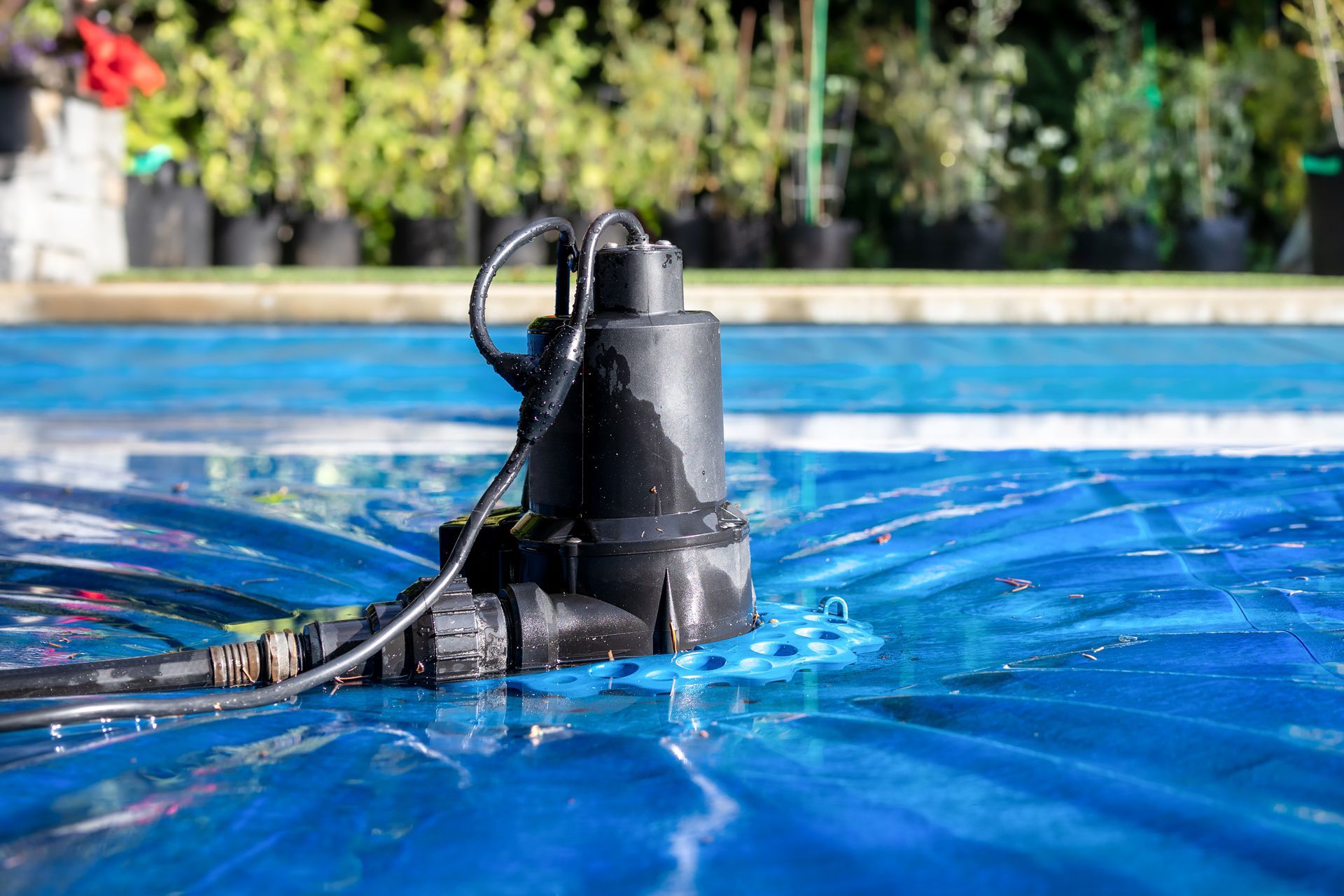 Black submersible pump on a blue pool cover, with a garden in the background.