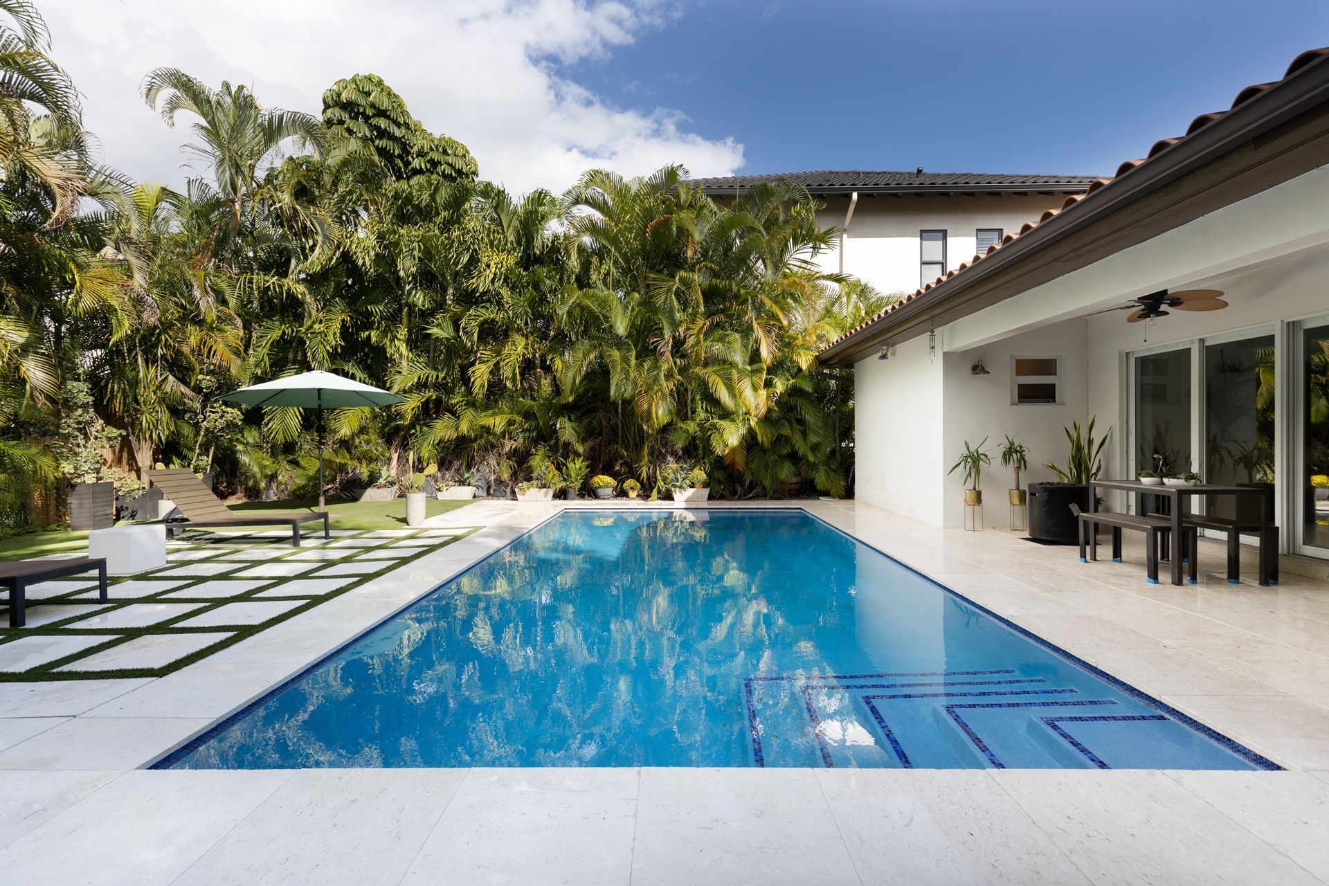 Pool with blue water and white tile, bordered by lawn and lush green.