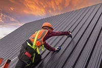 Person in safety gear on a metal roof, installing panels under an orange and purple sunset.