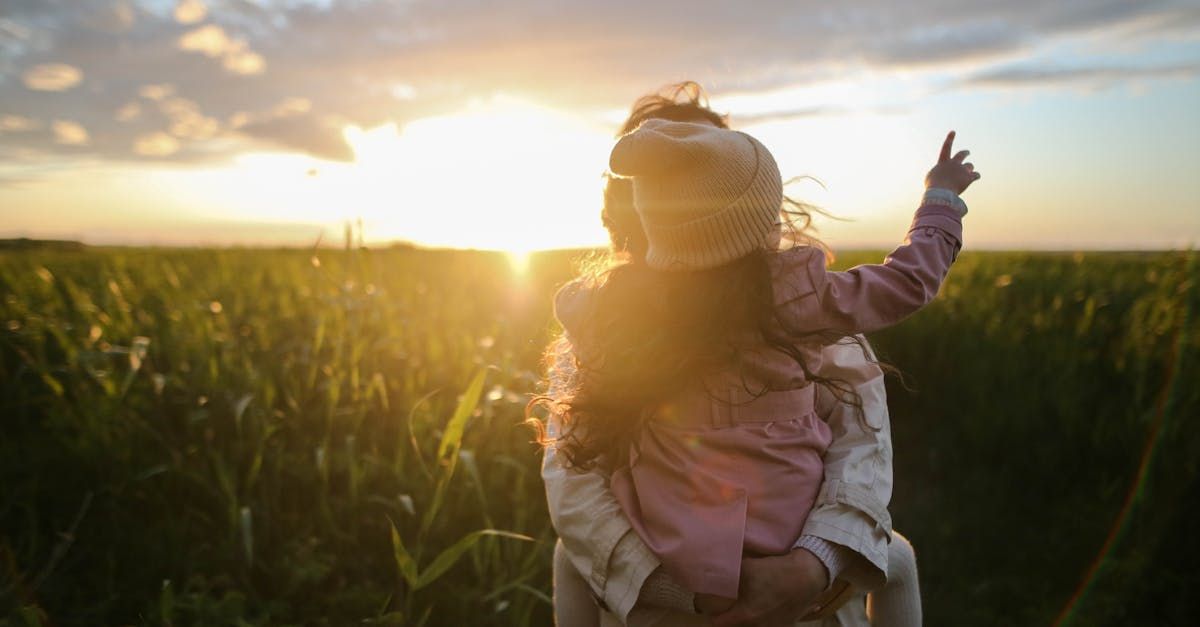 A woman is carrying a little girl on her shoulders in a field at sunset.