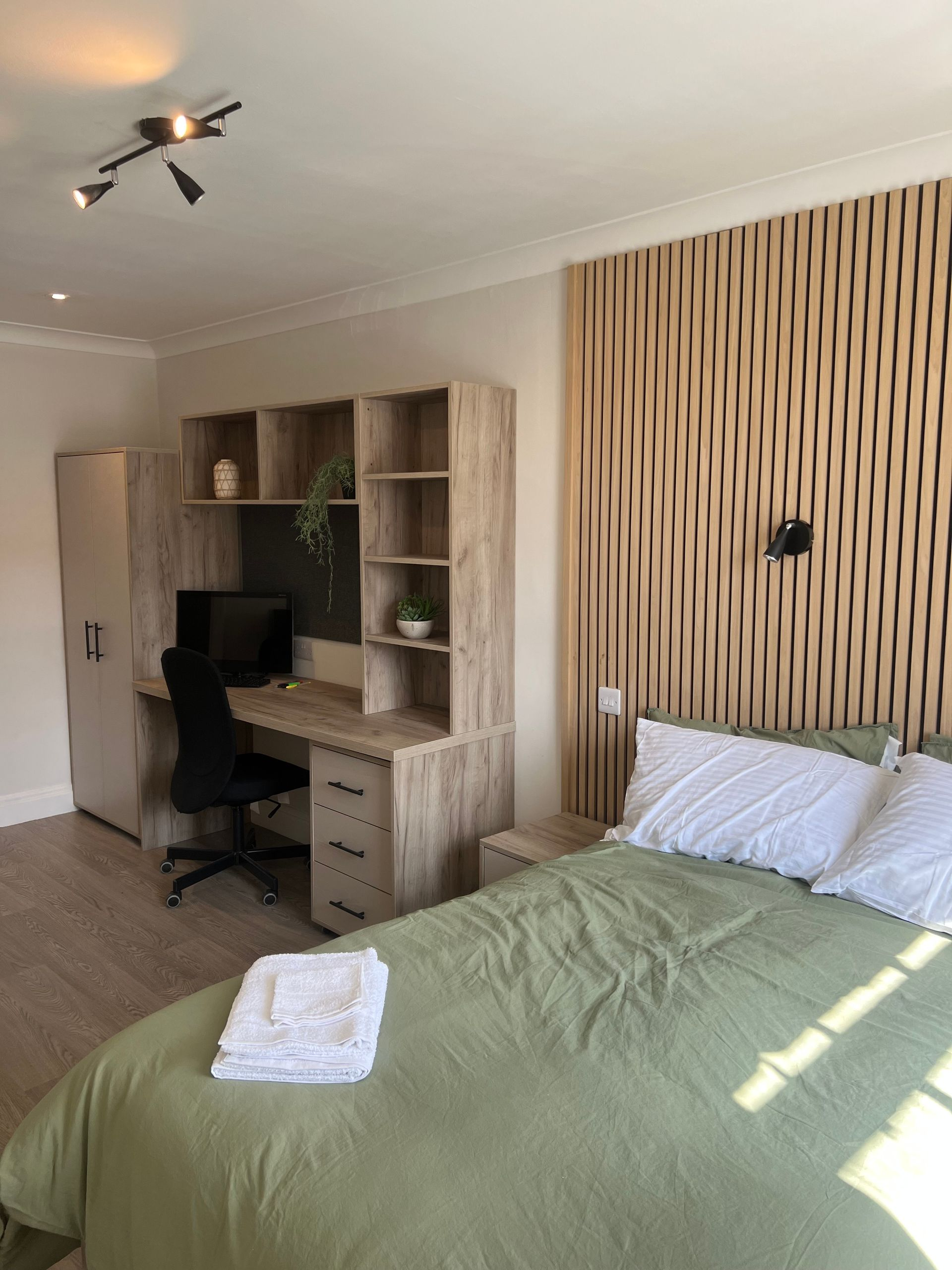 Bedroom with a desk and bed. Green bedding, wood accents on the wall, and a light-colored wooden desk.