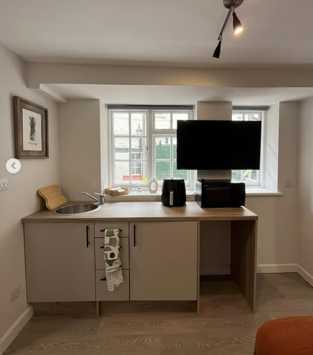 Small kitchenette with a sink, countertop, and TV above a small desk area, by a window.