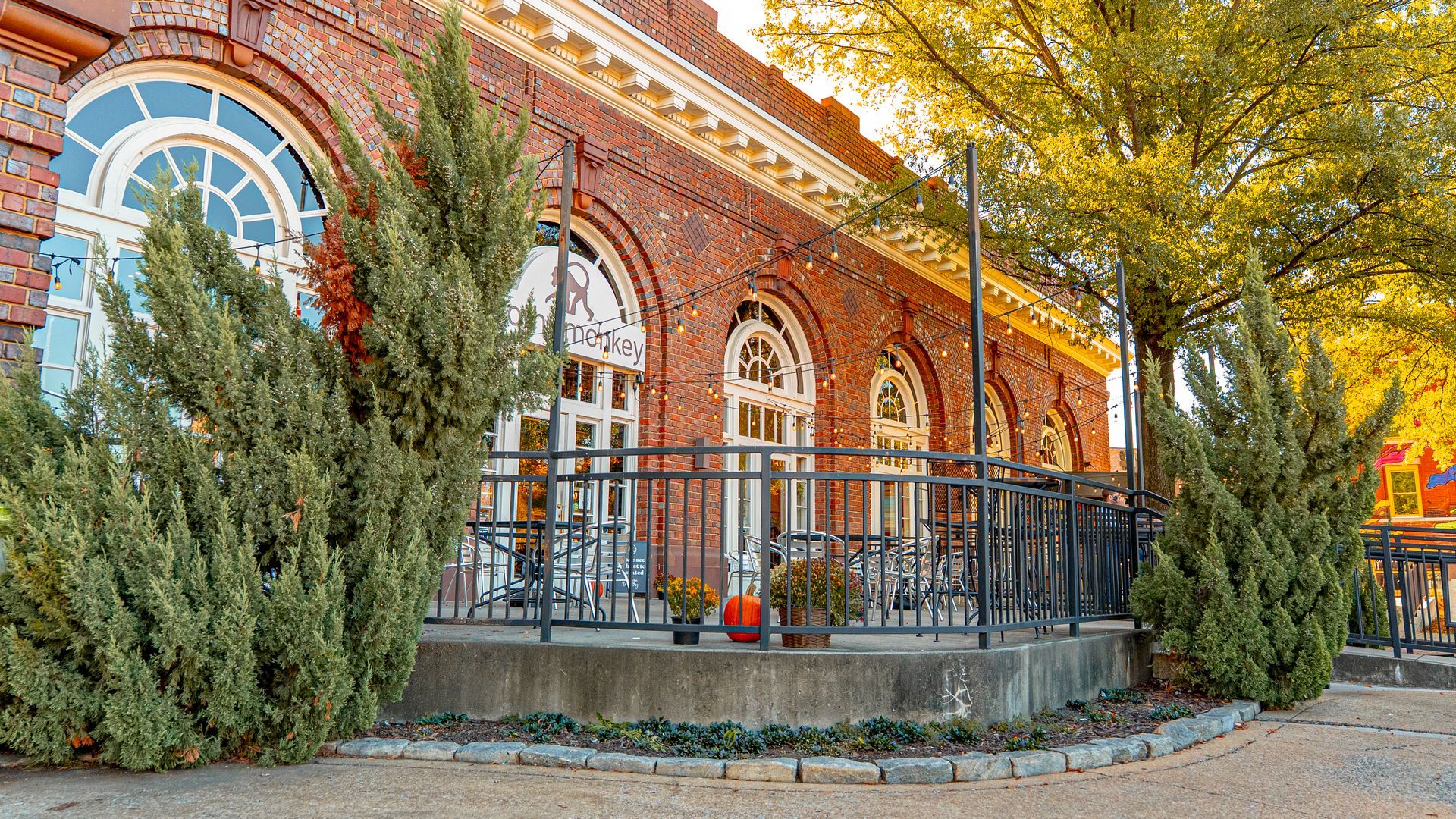Brick building with arched windows, outdoor seating on a patio. Green shrubs in front.