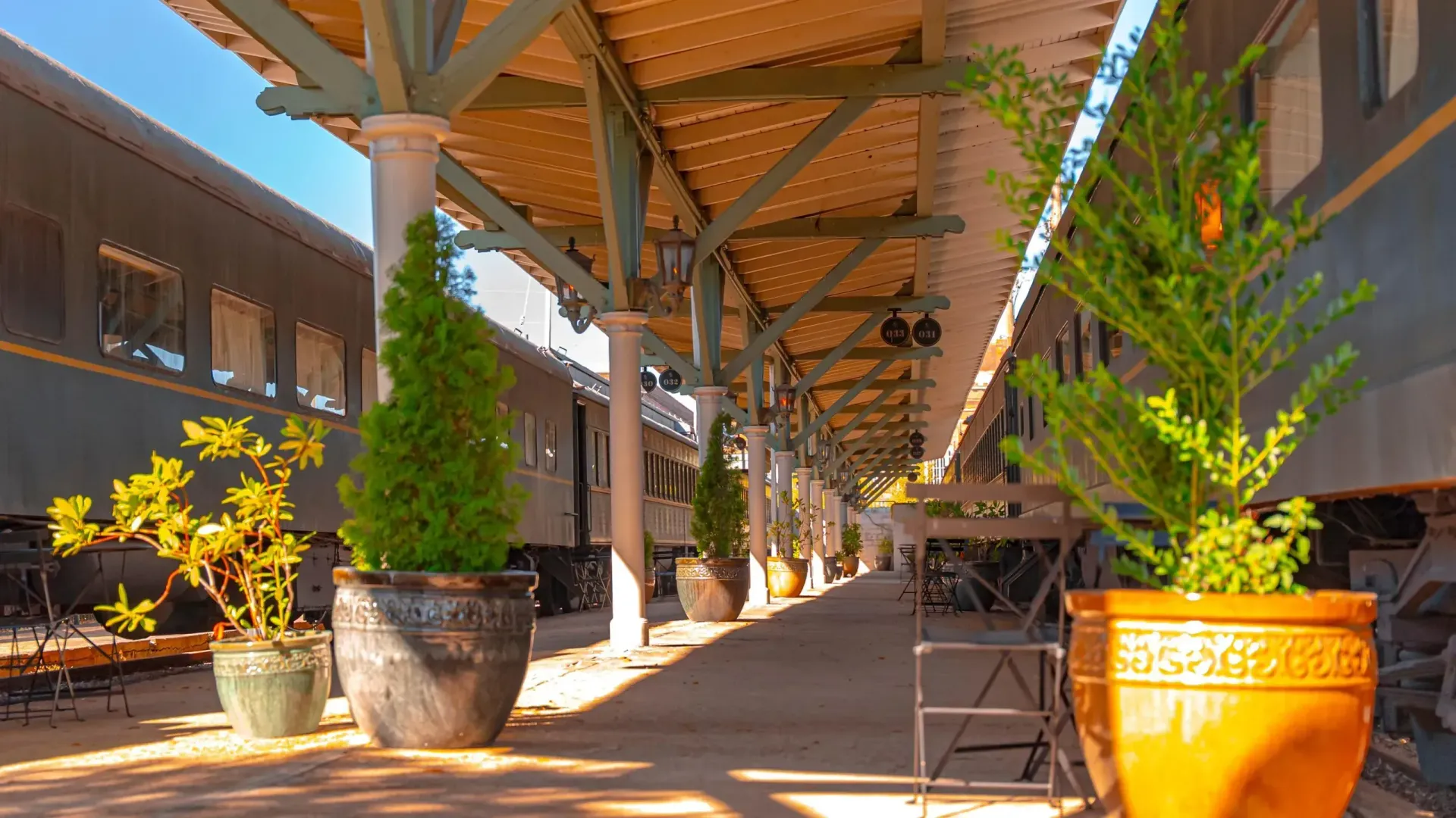 Train platform with covered walkway, potted plants, and vintage train cars.
