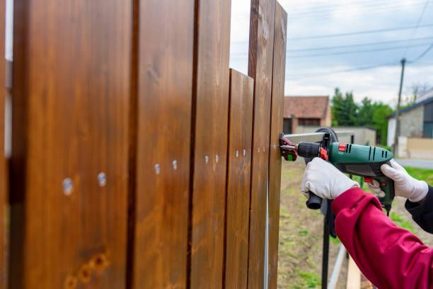 Person using a drill to attach boards of a stained wooden fence outdoors.