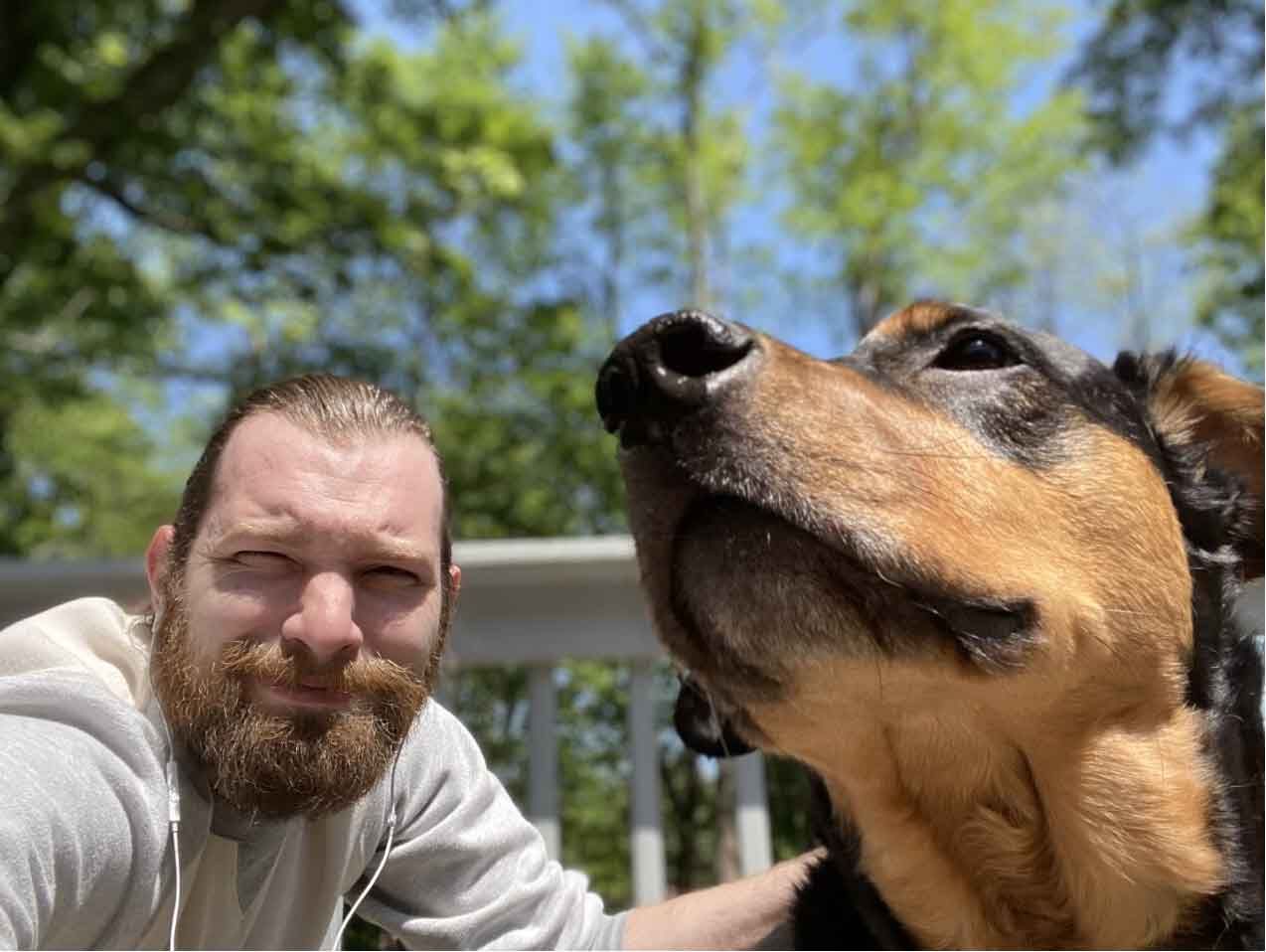 Man and dog posing for a selfie outside. Man has beard; dog has brown and black fur. Sunny, with trees in background.