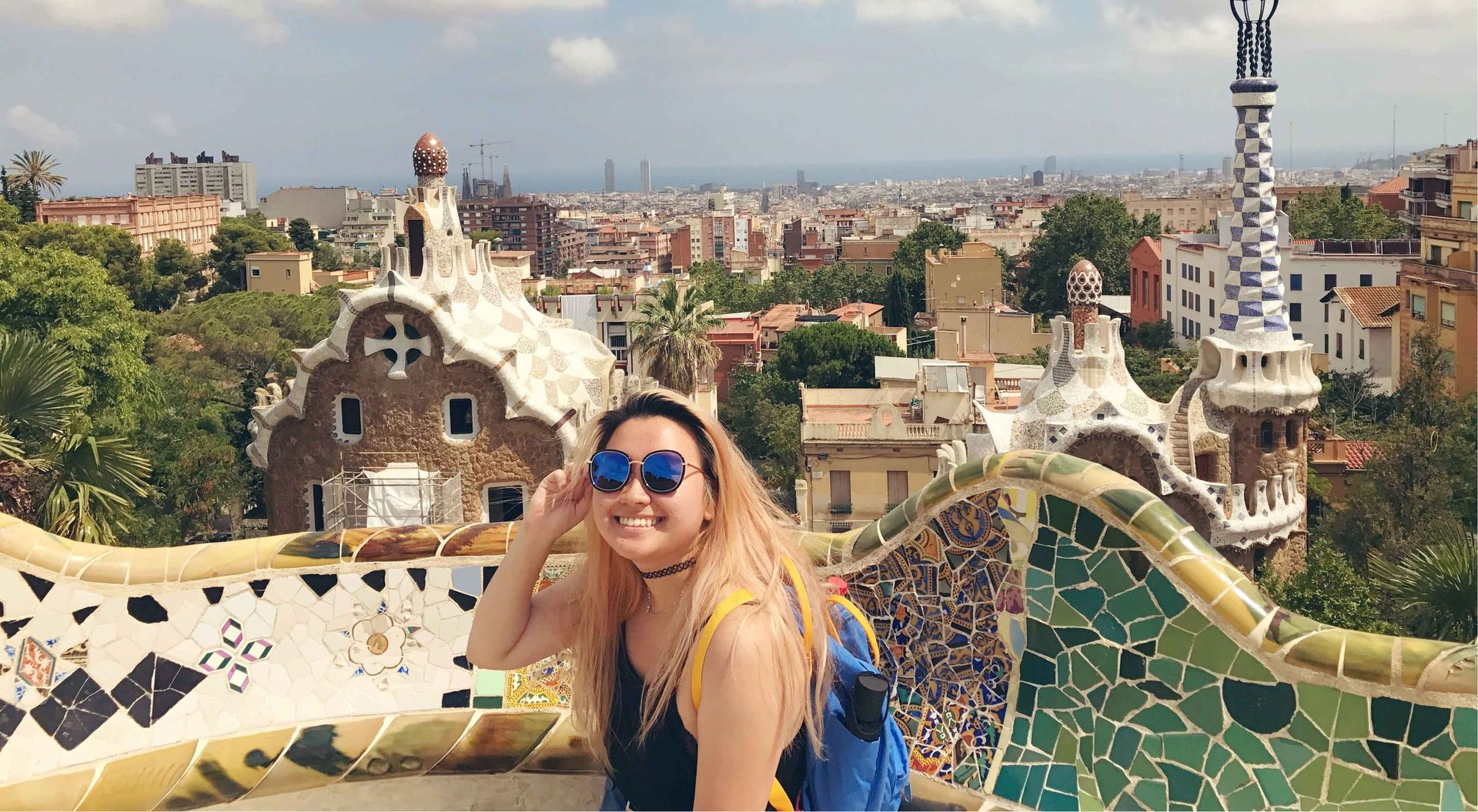 Woman smiling with sunglasses, posing at Park Güell, Barcelona, colorful mosaic bench in foreground, city view.