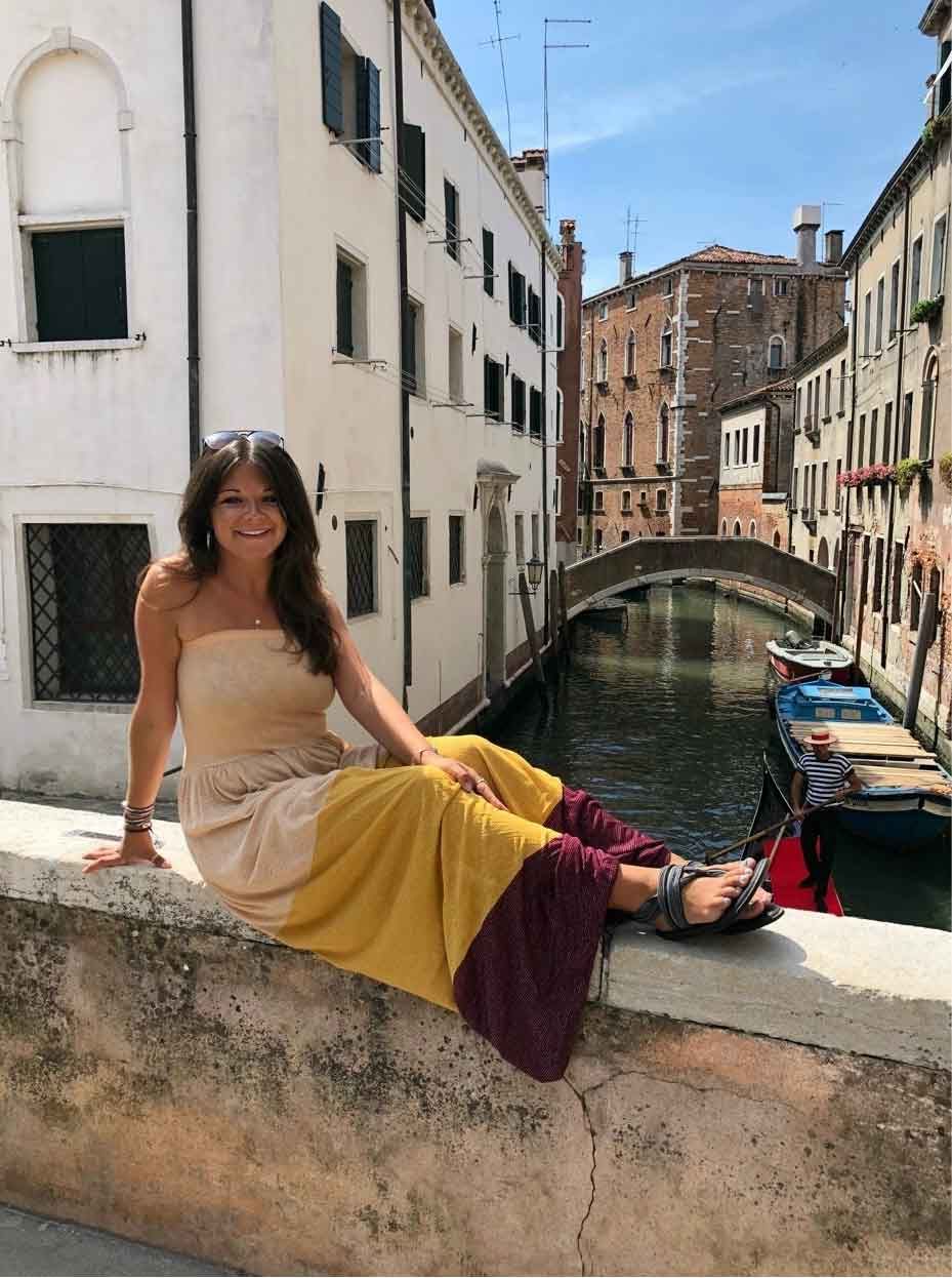 Woman sitting on a stone wall in Venice, Italy, overlooking a canal with a bridge and buildings.