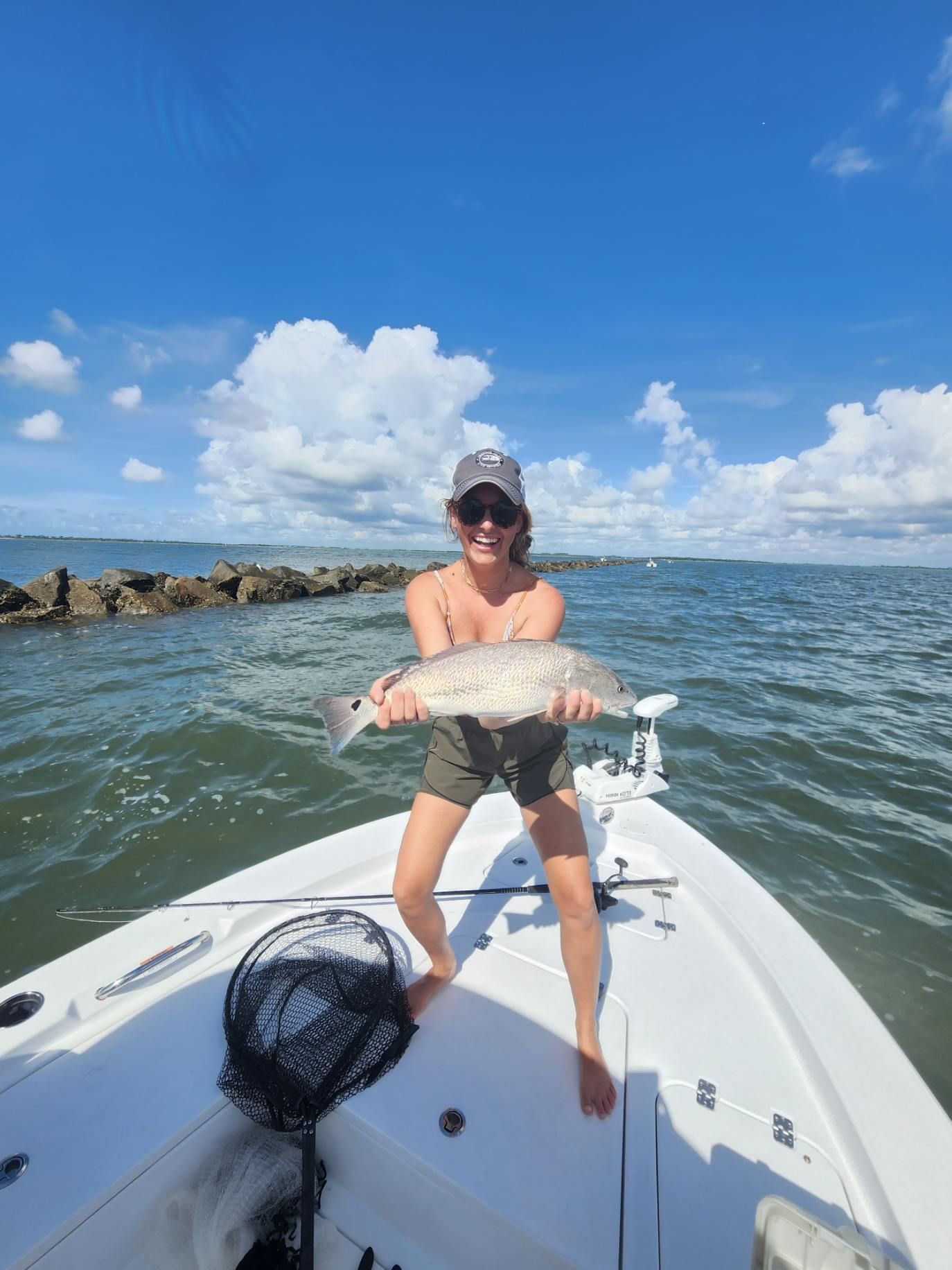 Woman on boat holding up large fish, blue sky, water, jetty in background.