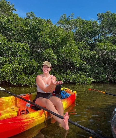 Woman kayaking, smiling, holding something. Kayak is red/yellow, water and trees behind her.