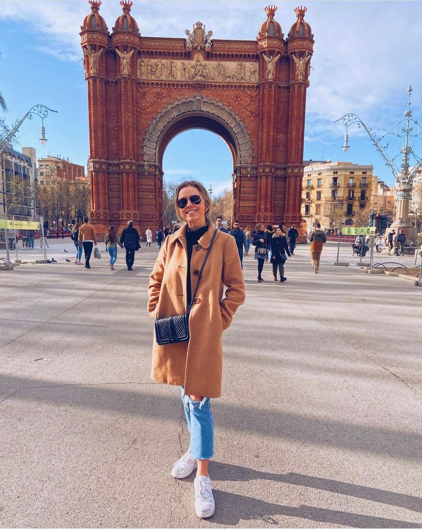 Woman in a tan coat and jeans stands in front of the Arc de Triomf in Barcelona.