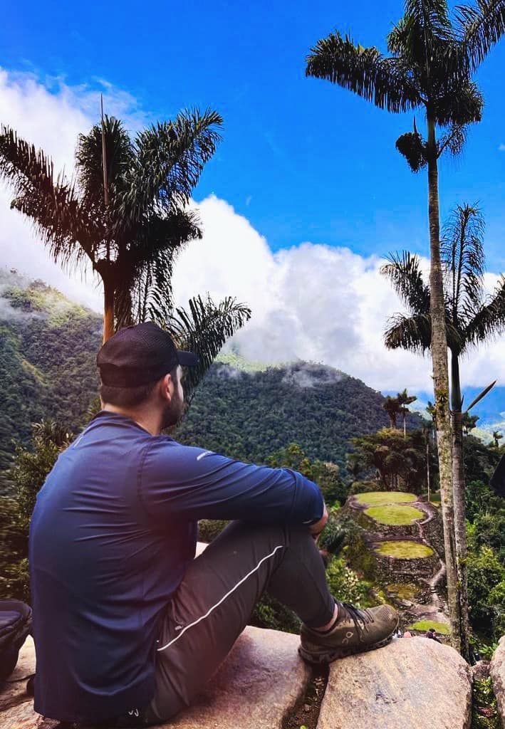 Man in blue shirt, sitting, looking at mountains with trees and cloudy blue sky.