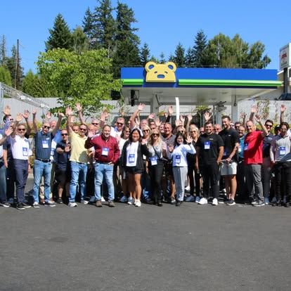 Group of people waving in front of a gas station with a bear logo under a blue sky.