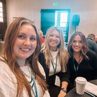 Three women smiling at a conference, one holding a coffee cup, indoor setting.