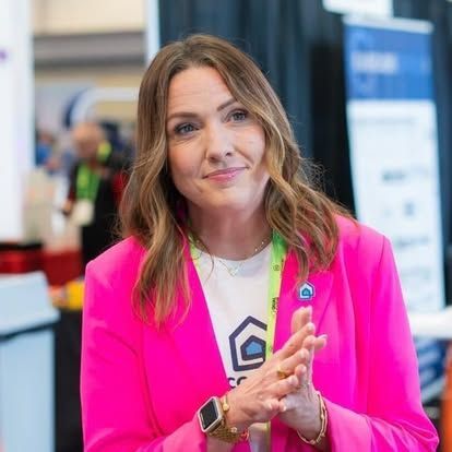 Woman in a pink blazer at a conference, hands clasped. She is wearing a white shirt and watch.