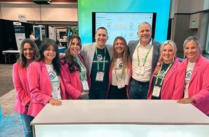 Group of people at a booth smiling; wearing pink blazers, green accents, and name tags; standing behind a table.
