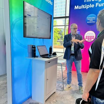 Man gestures towards a screen while speaking to another person at a conference booth. Blue/green backdrop.