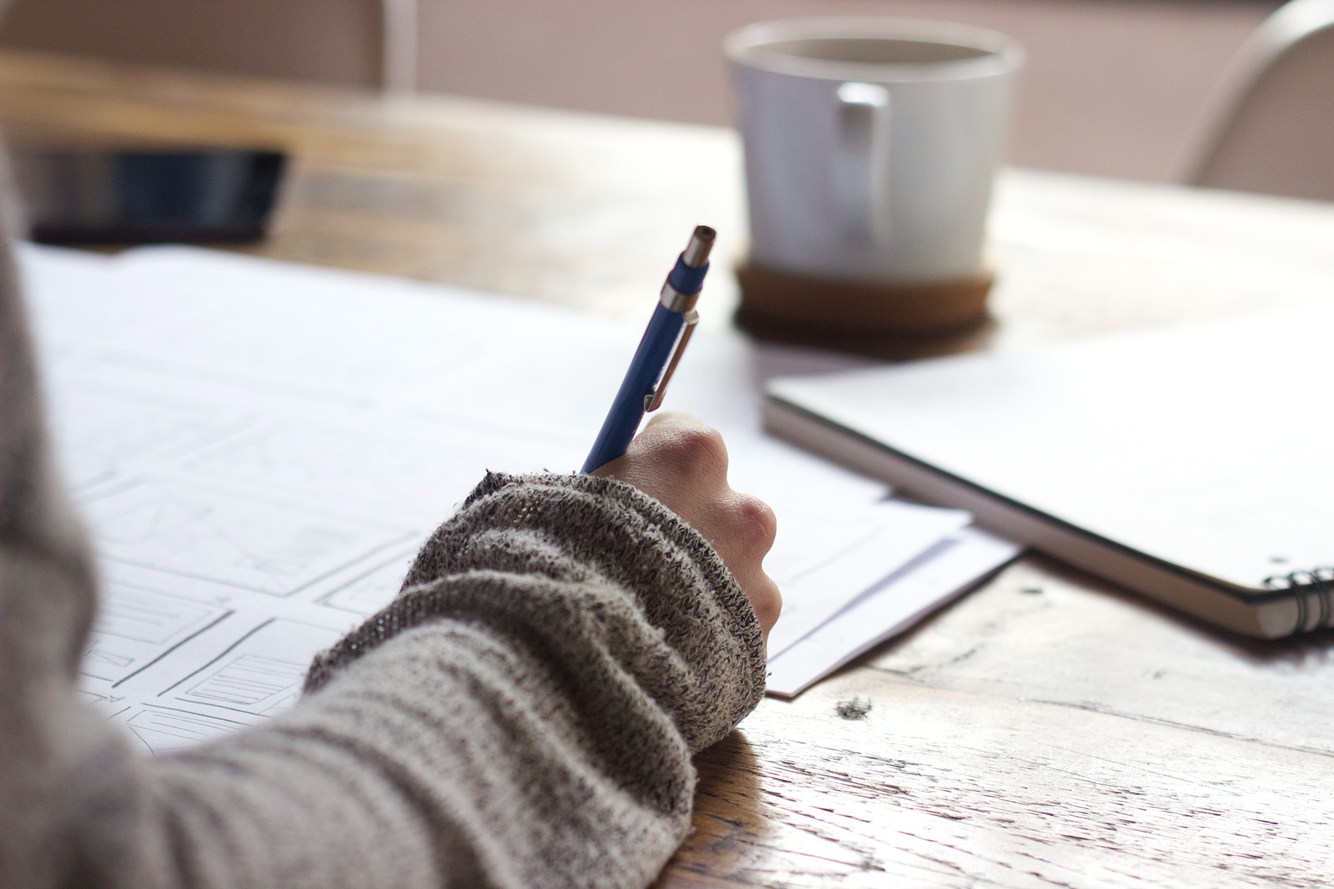 A person in a textured sweater holds a blue mechanical pencil while writing on papers at a wooden table with a coffee mug.