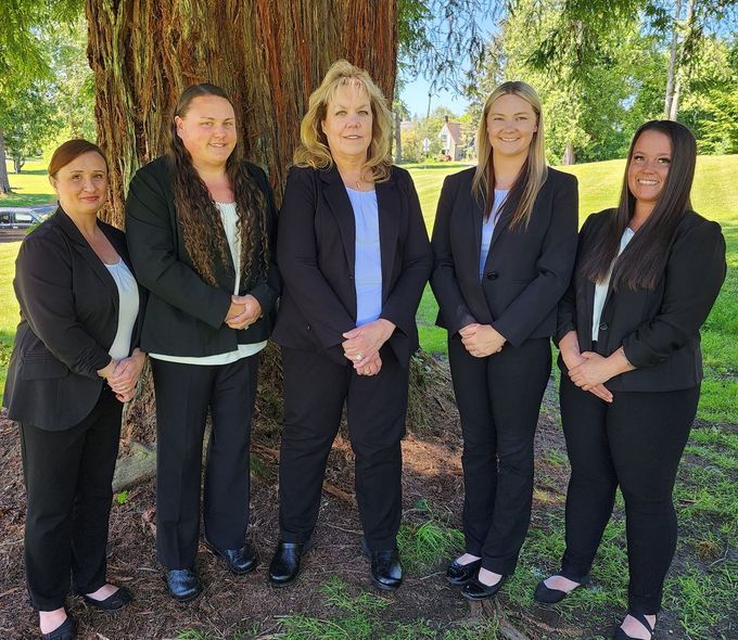 Five people in professional business attire stand in a row outdoors in front of a large tree.
