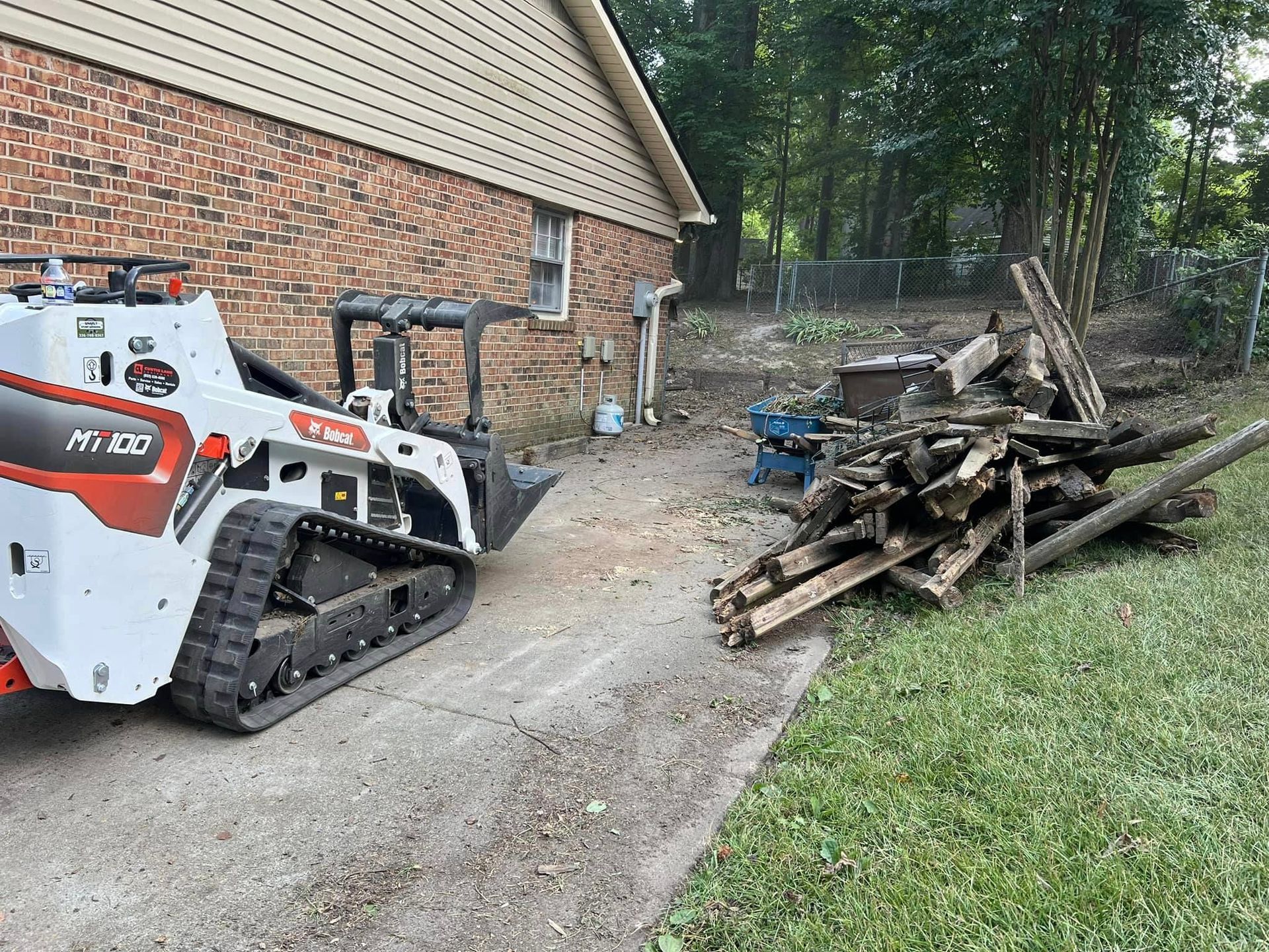 A bulldozer is parked in front of a brick house next to a pile of wood.
