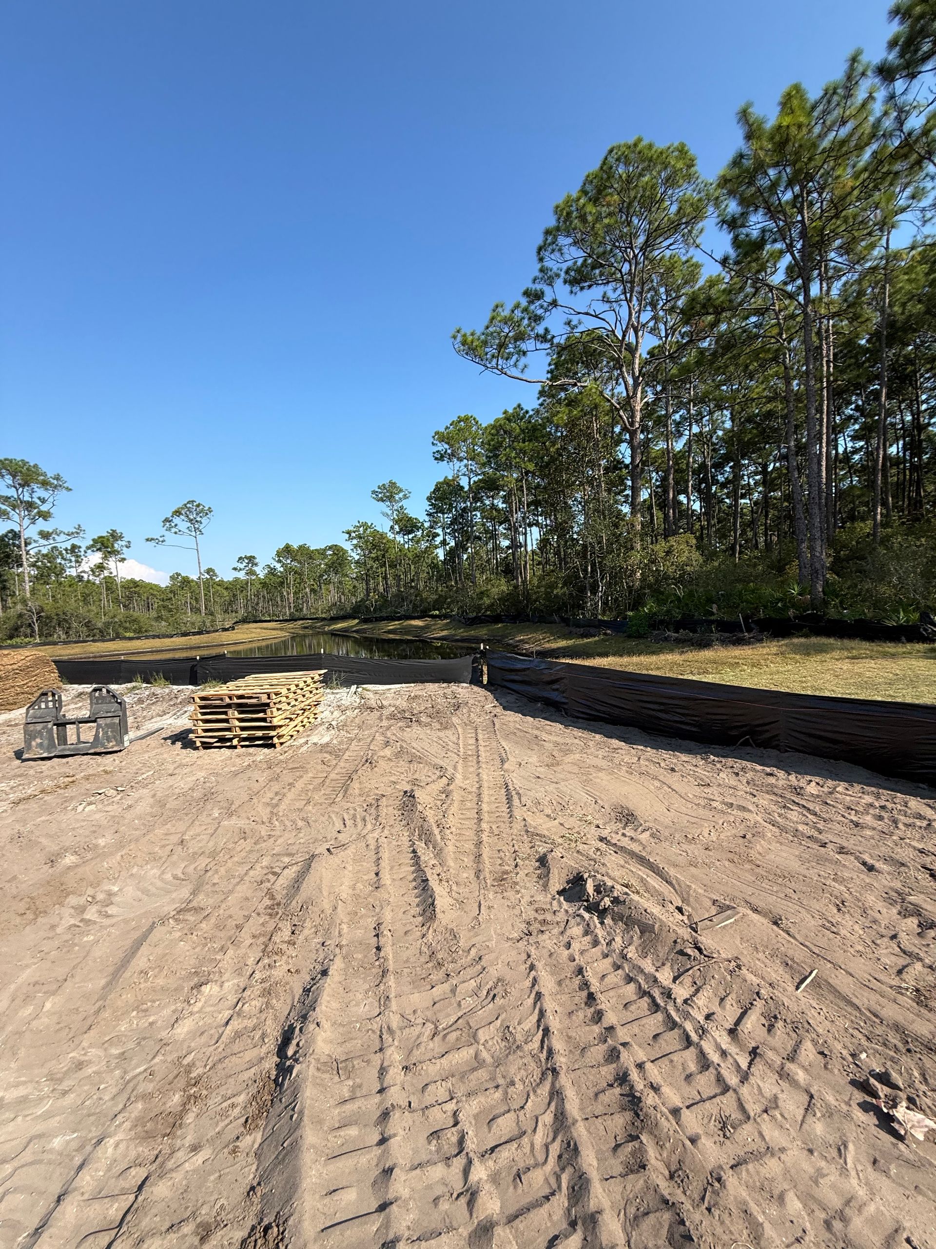 Dirt lot with tire tracks and construction materials, with trees and a blue sky in the background.