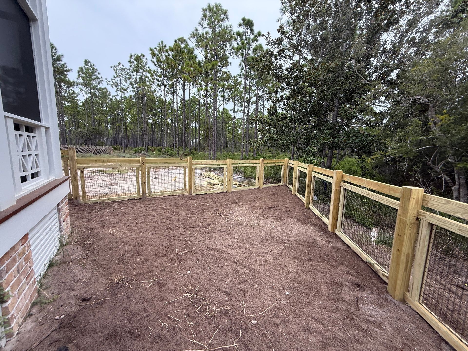 A newly built wooden fence encloses a dirt patch with a forest background next to a house.
