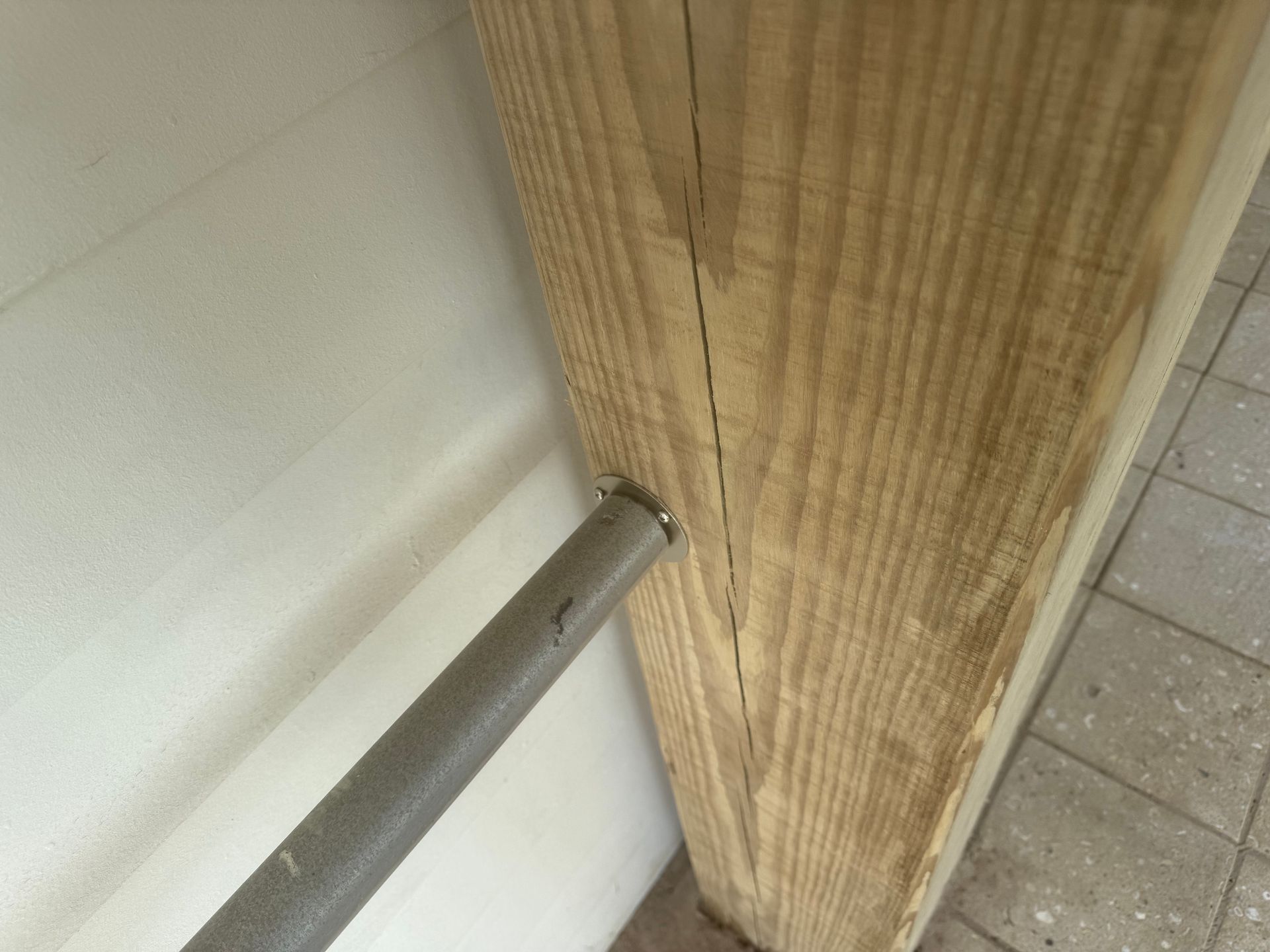 A metal bar piercing a wooden beam, near a white wall and tiled floor.