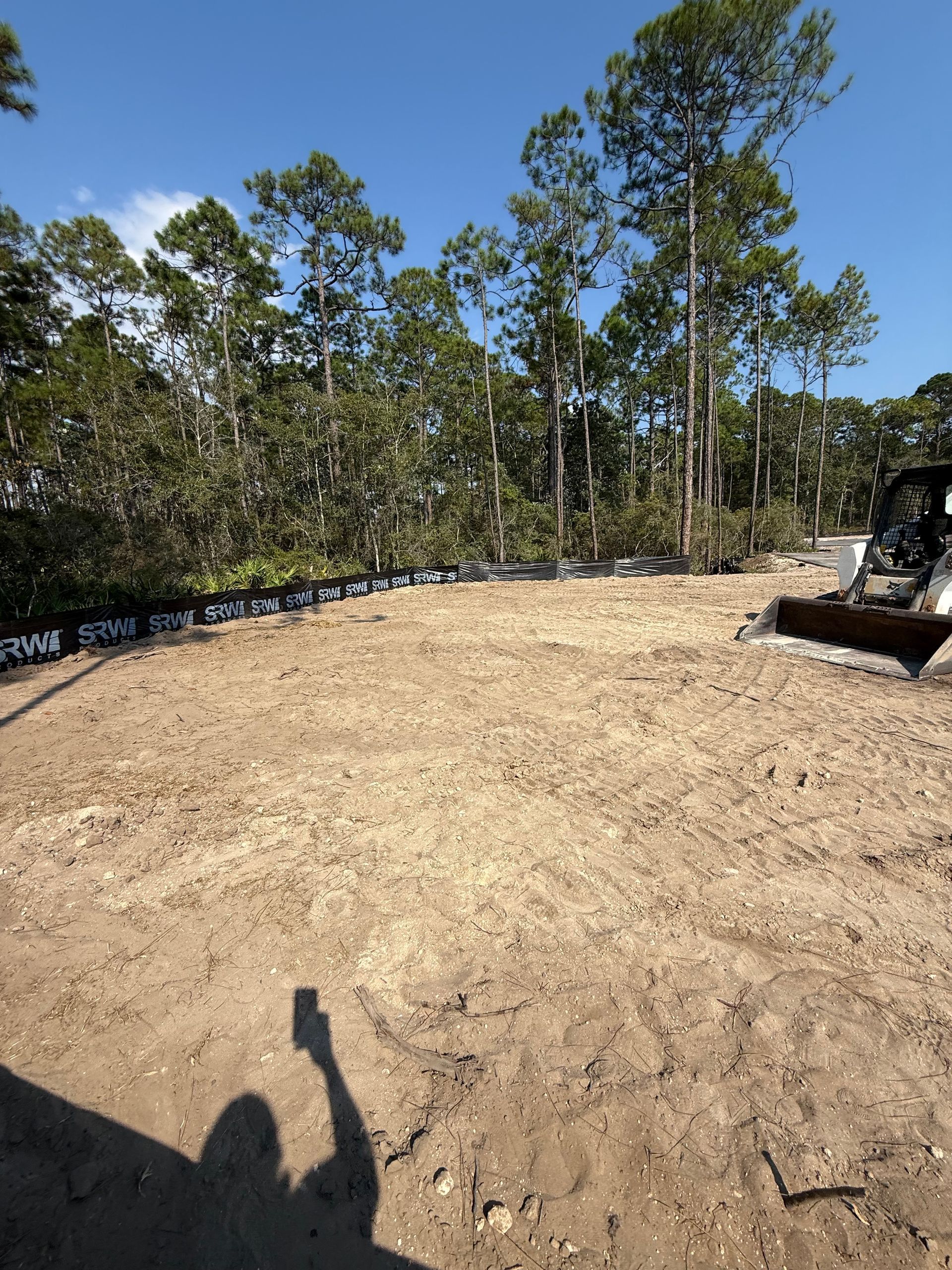 Cleared dirt lot with trees in the background. A shadow of a person takes a photo.