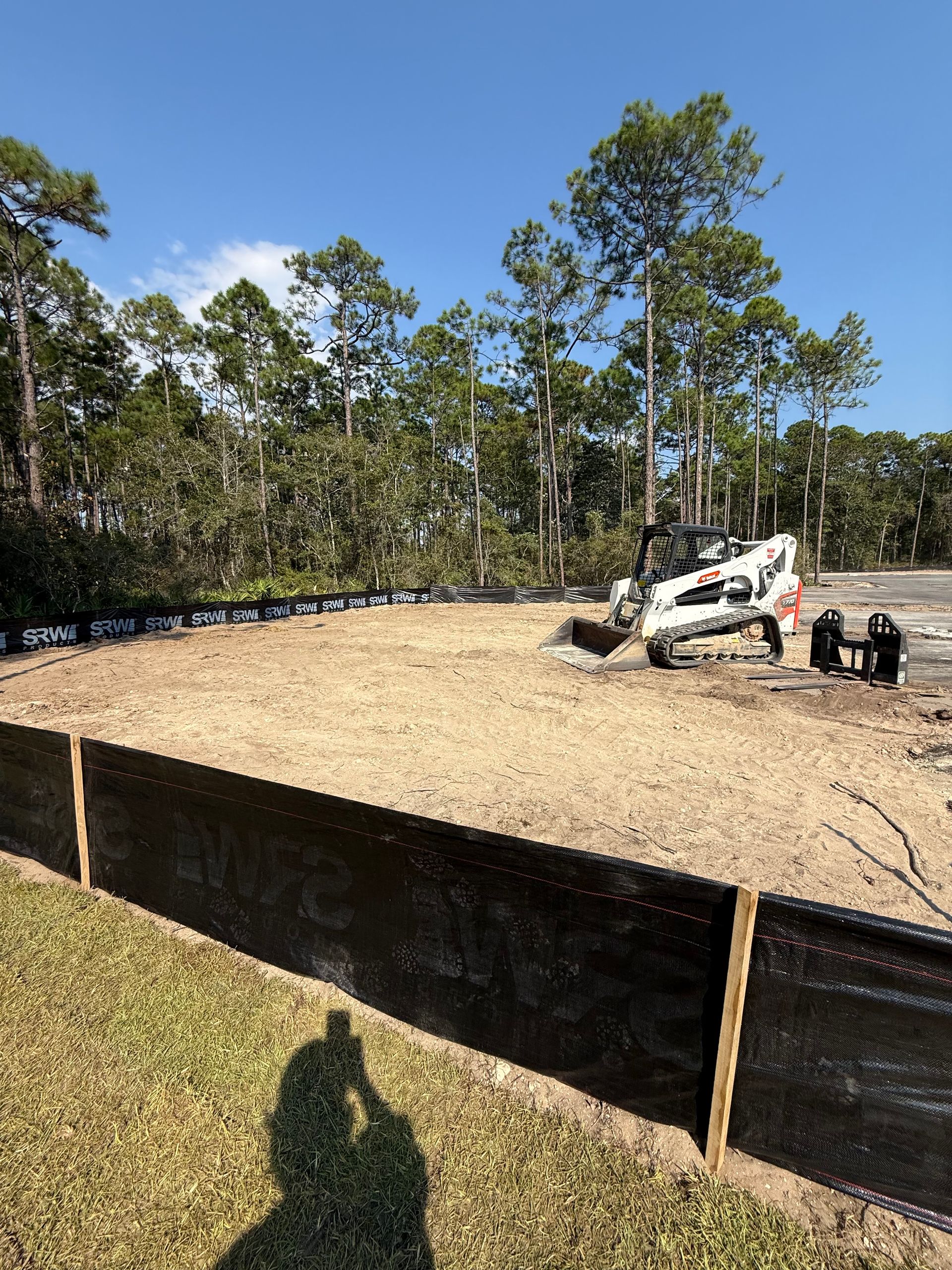 Construction site with black silt fence, dirt, trees, and a skid steer on a sunny day.