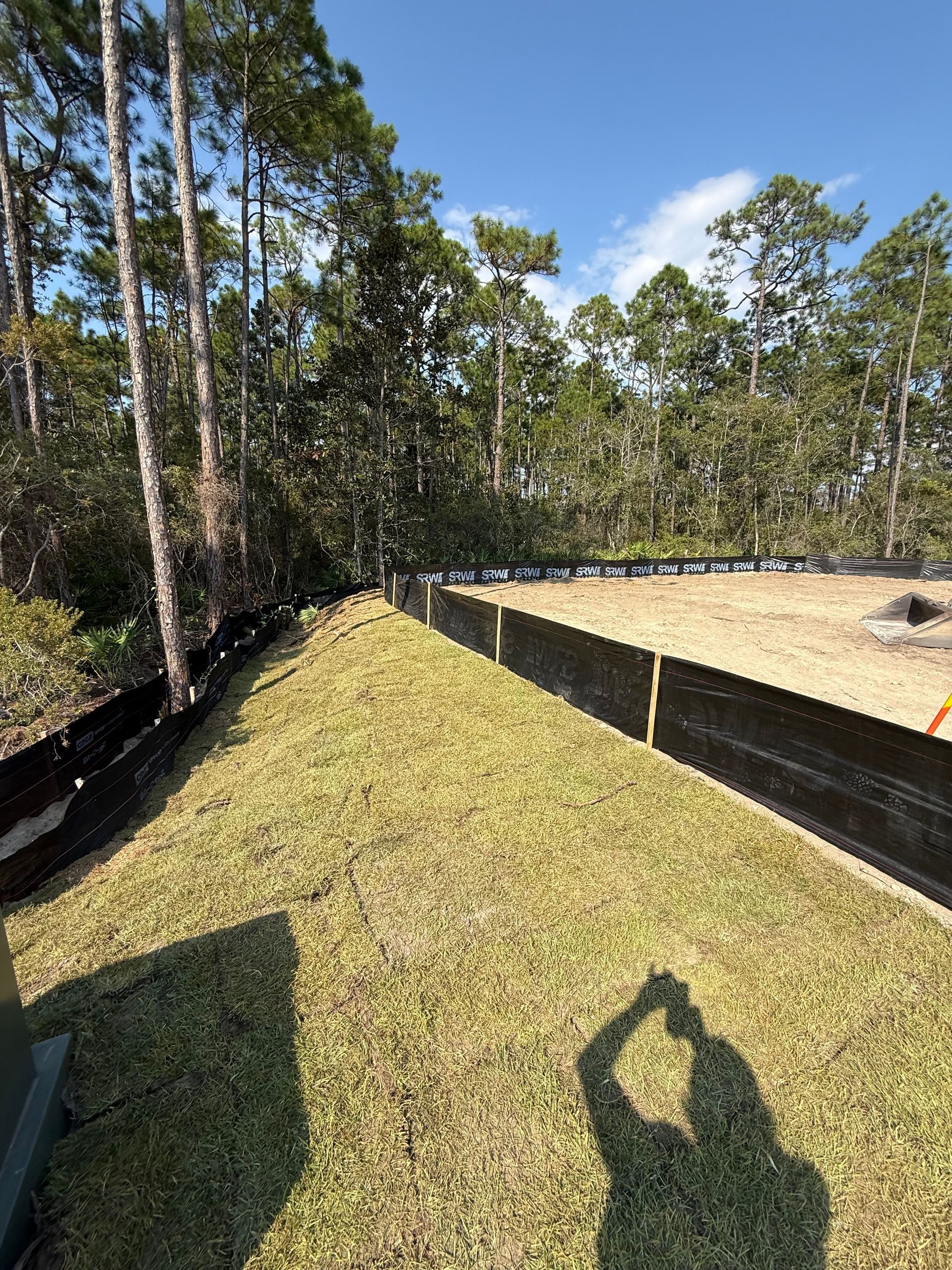 A grassy area with a black barrier fence, trees in the background, and a shadow in the foreground.