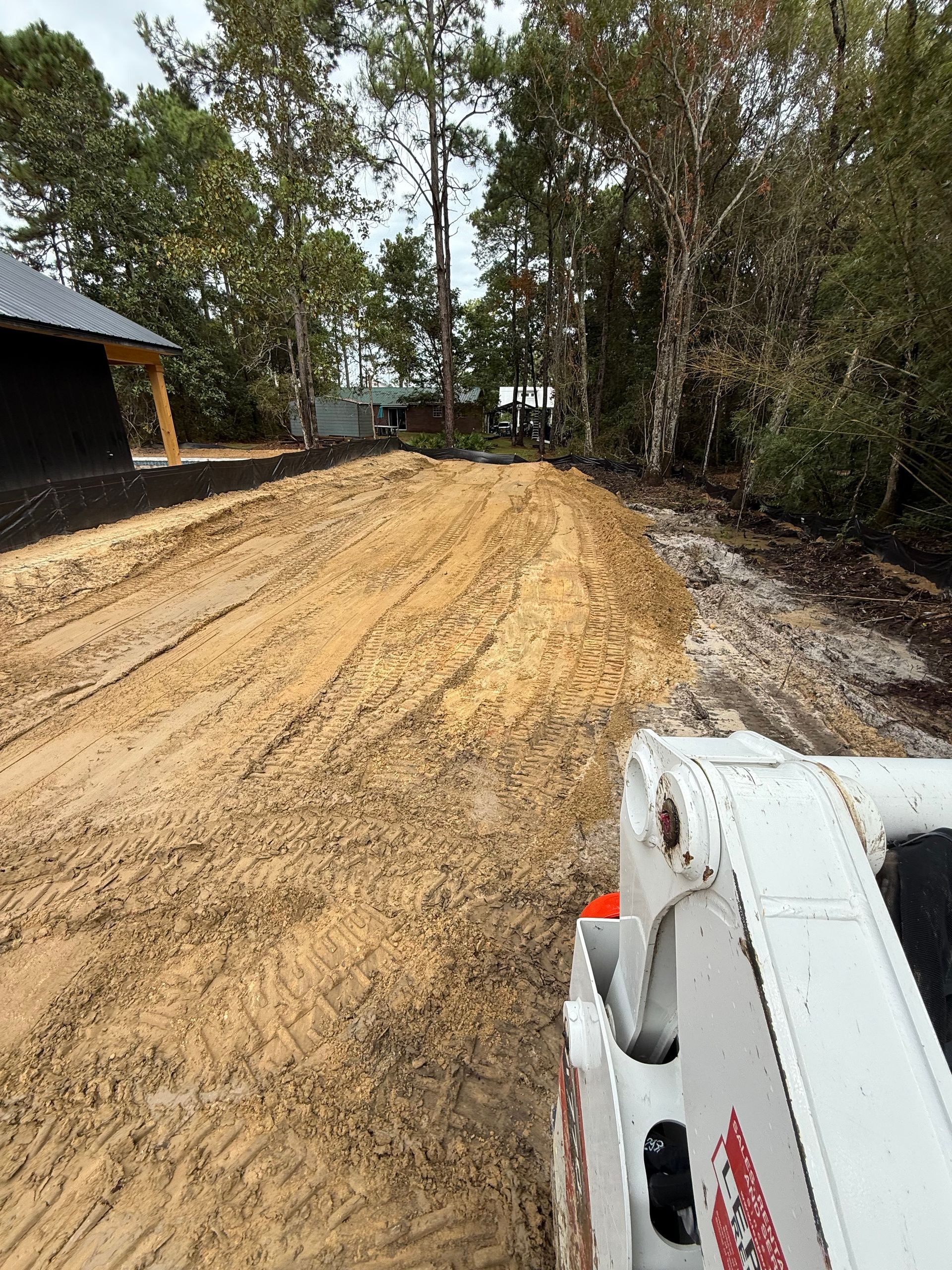Muddy construction site with a skid steer in the foreground; trees and a building in the background.