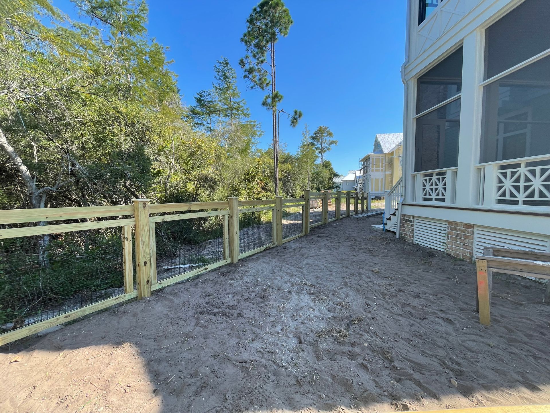 New wooden fence along a dirt path next to a house with a screened porch, under a blue sky.