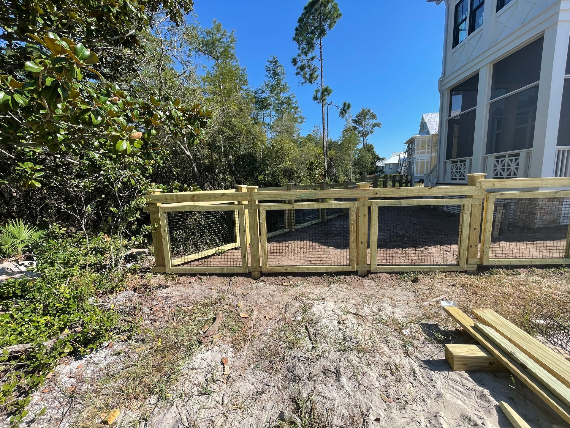 A newly constructed wooden fence surrounds an area with brush and small trees. Buildings visible in the background.