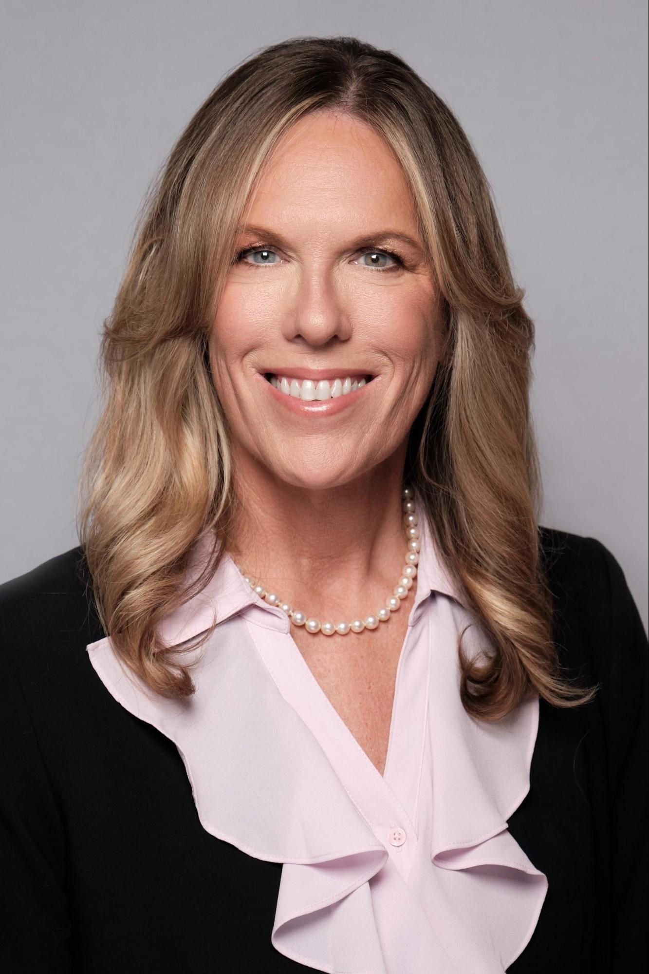 A woman wearing a pink blouse and a pearl necklace is smiling for the camera.