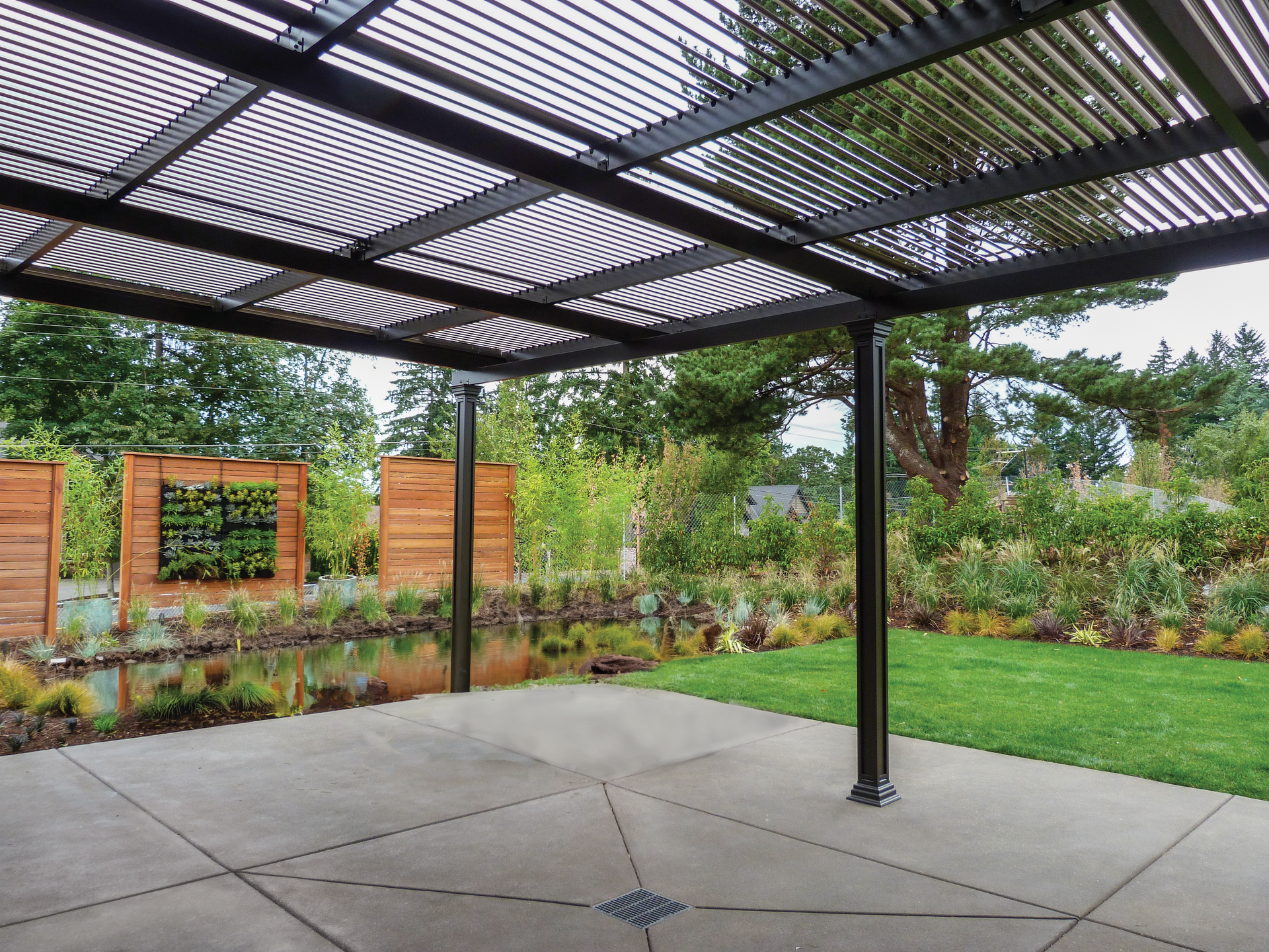 Pergola with a concrete patio, overlooking a garden with a pond, and a wooden fence.