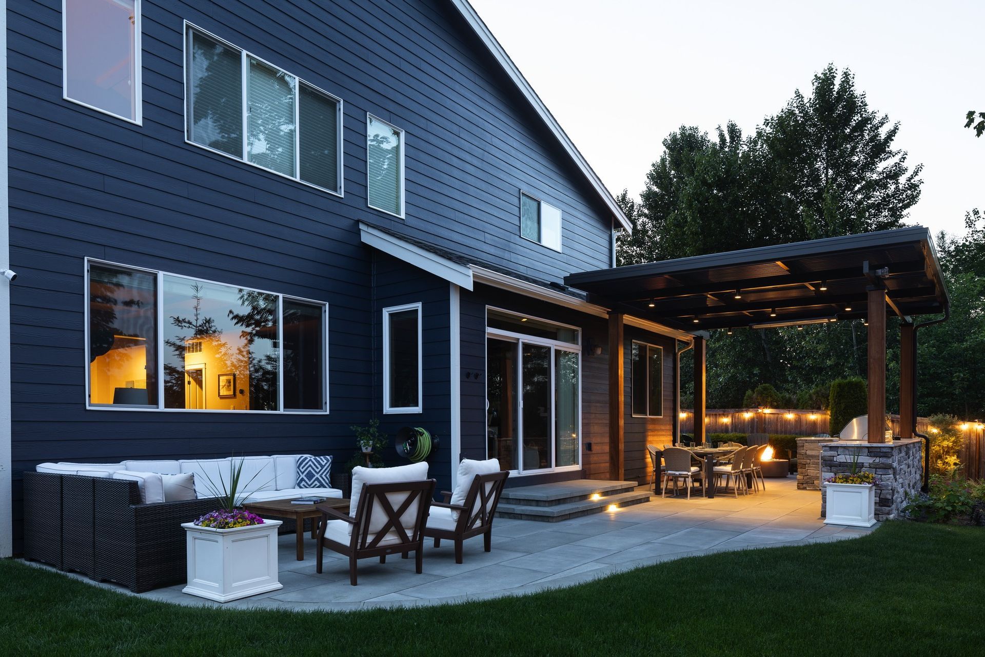 A two-story blue house with an illuminated patio, furniture, and string lights at dusk.