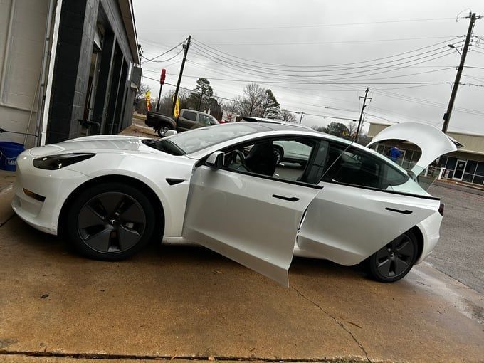 White Tesla Model 3 with open doors parked outside a building on a wet, overcast day.