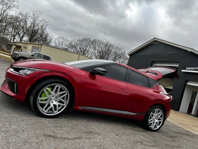 Red Kia EV6 with silver wheels, tinted windows, and open trunk, parked outside a building on a cloudy day.
